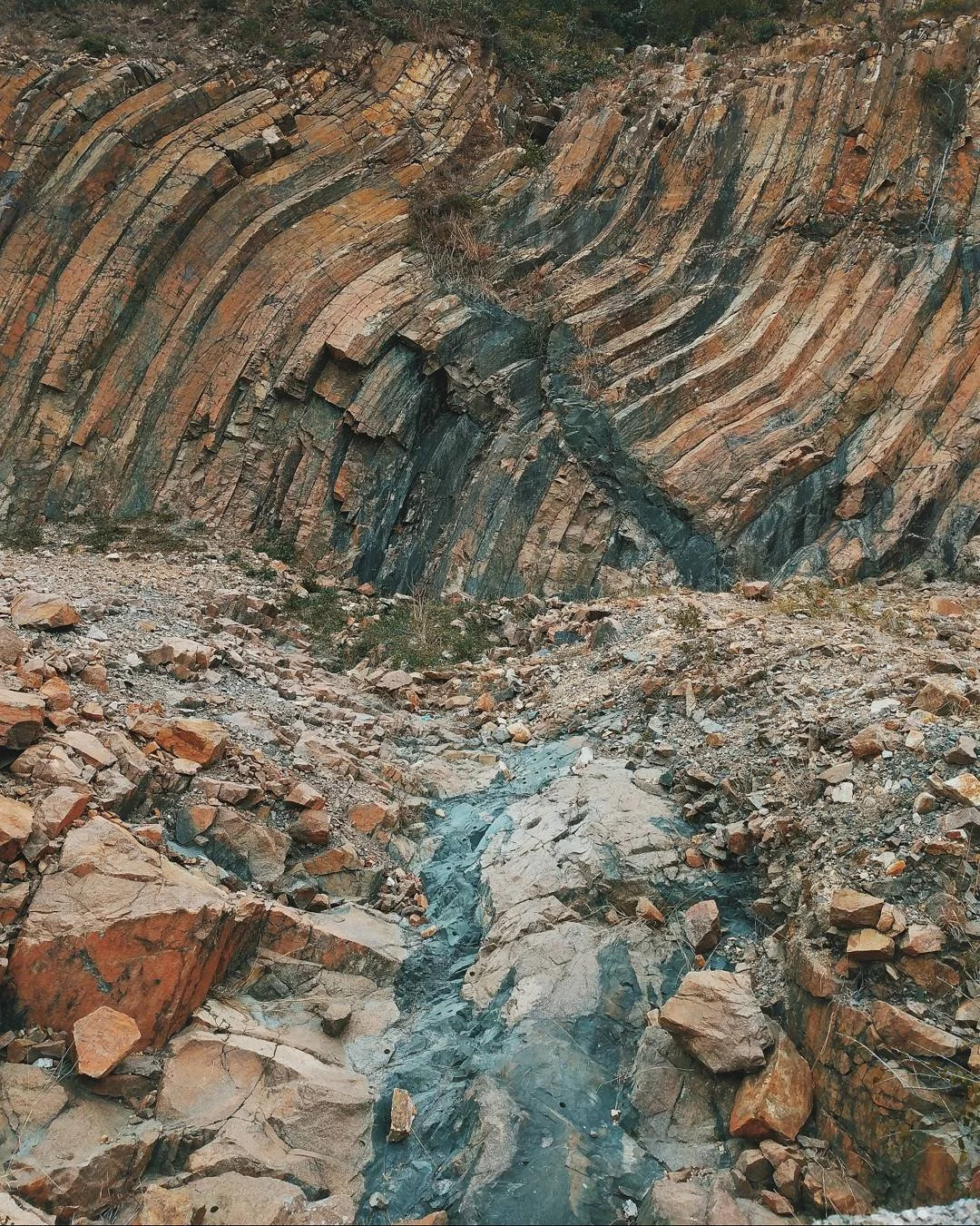 Rocky cliff with layered, tilted strata and small water stream at the base.