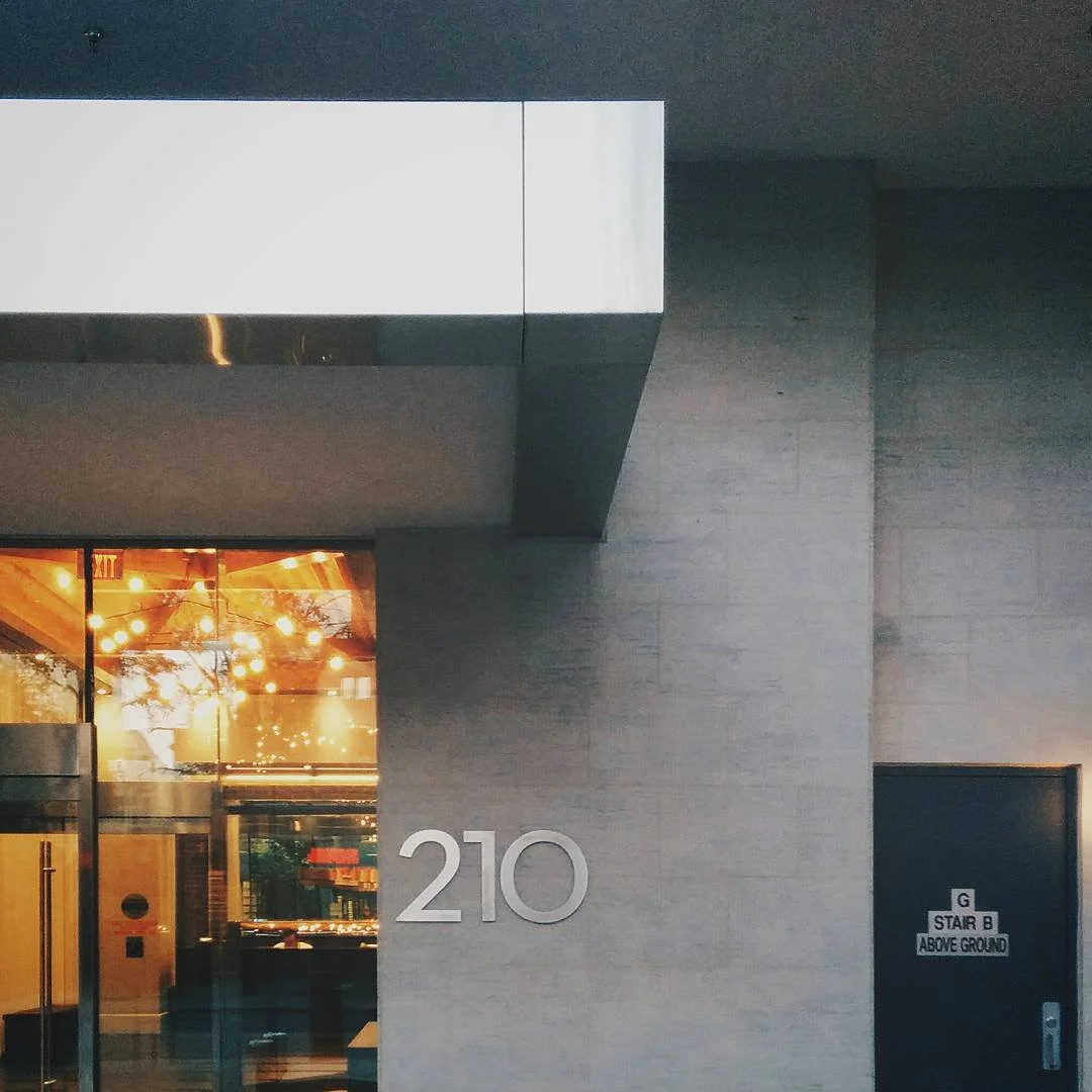 Exterior of a building with the number 210 on the wall. There is a glass door reflecting warm lights and interior space. A sign on a door indicates 'STAIR B ABOVE GROUND'.