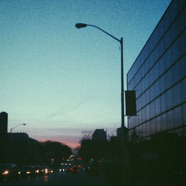 City street scene during twilight with streetlights, cars with headlights, dark silhouettes of buildings, and a large modern building on the right.
