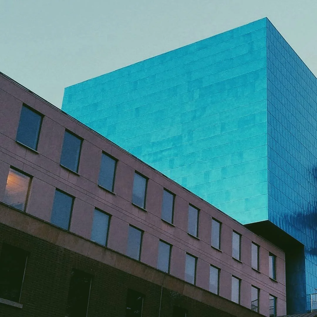 A modern cityscape with a large blue glass office building towering above a smaller beige building with rectangular windows.