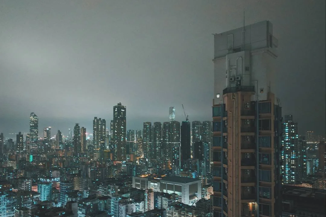 Nighttime cityscape of a dense urban area with high-rise buildings and skyscrapers, some with illuminated windows, under a cloudy sky.