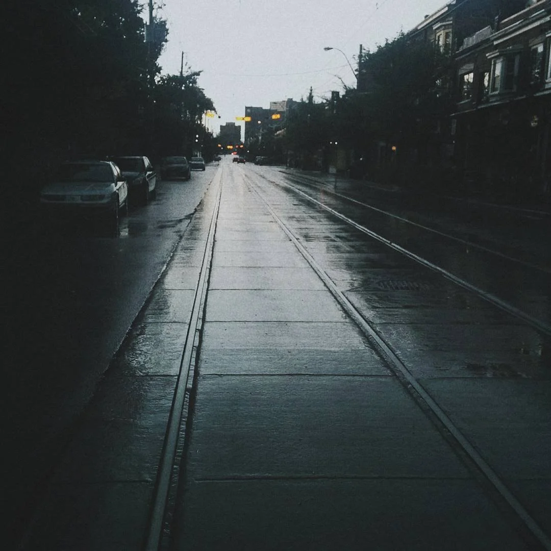 Rainy city street with wet pavement and parked cars on the left, overhead tram tracks, buildings on the right, and traffic lights in the distance.