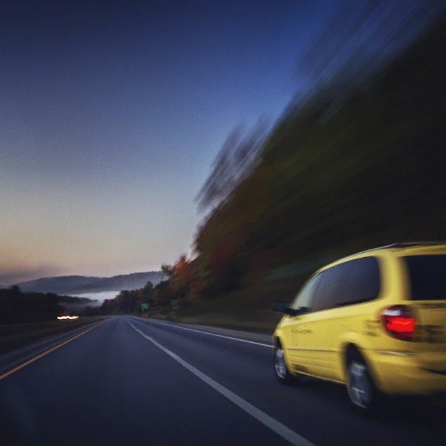 A yellow minivan driving on an open highway during dusk with motion blur effects.
