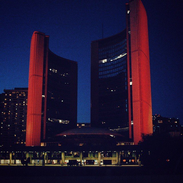 Nighttime photo of a modern, tall building with curved, dark glass and red vertical accents, illuminated by nearby lights, with a clear sky in the background.