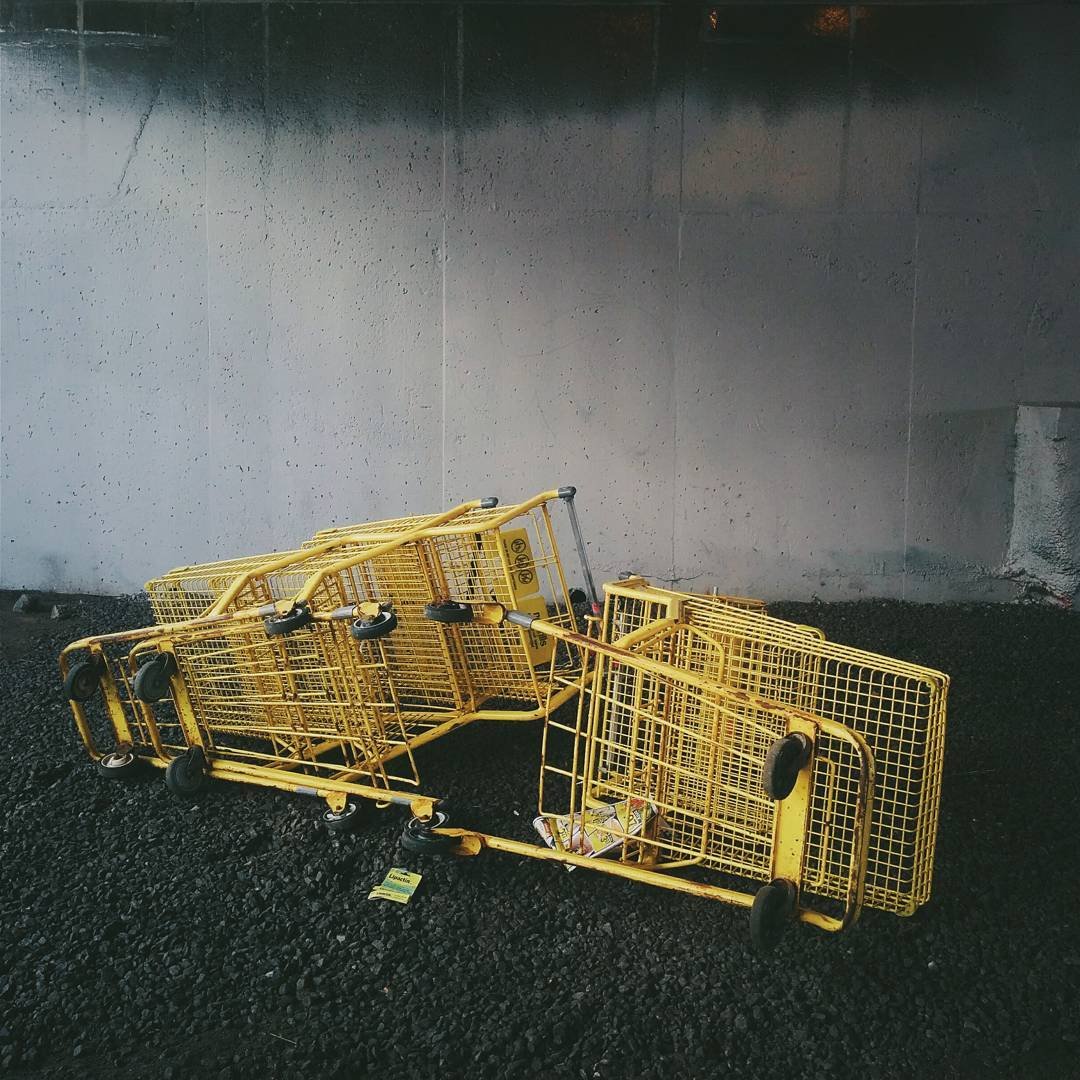 Several yellow shopping carts are tipped over and piled on a dark asphalt ground against a concrete wall.