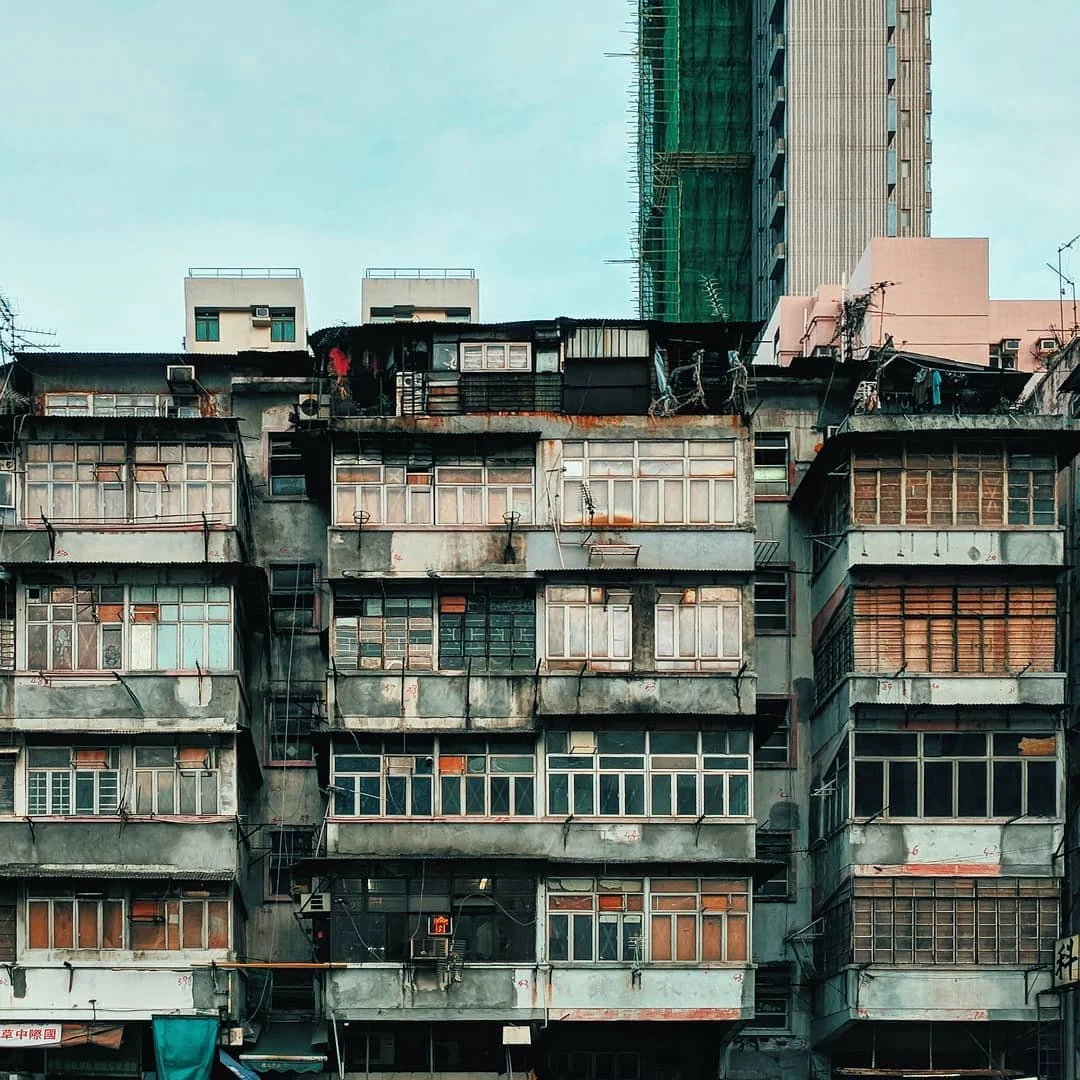 Old, weathered apartment building with multiple windows and balconies, some with rusted metal railings, in front of modern high-rise buildings under construction.