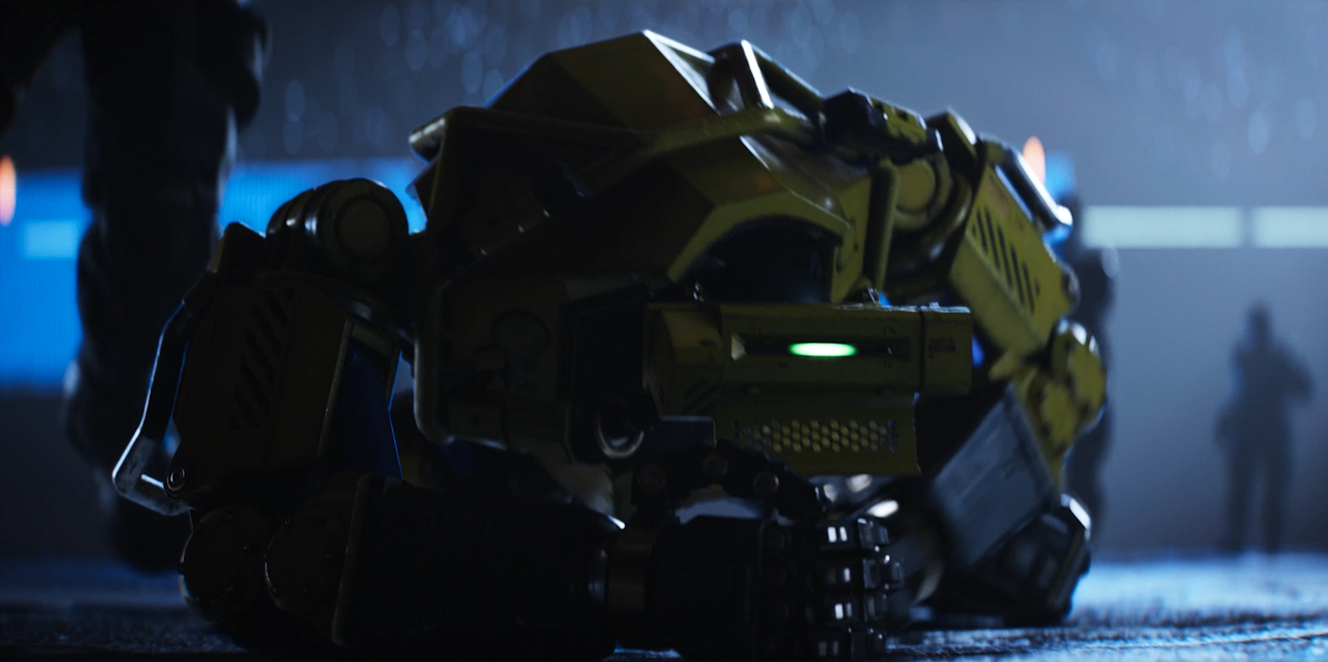 A yellow futuristic robot or vehicle on the ground in a dark blue-lit setting, with silhouetted figures in the background.