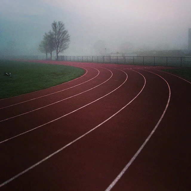 An outdoor running track on foggy day with empty lanes, trees, and a fence in the background.