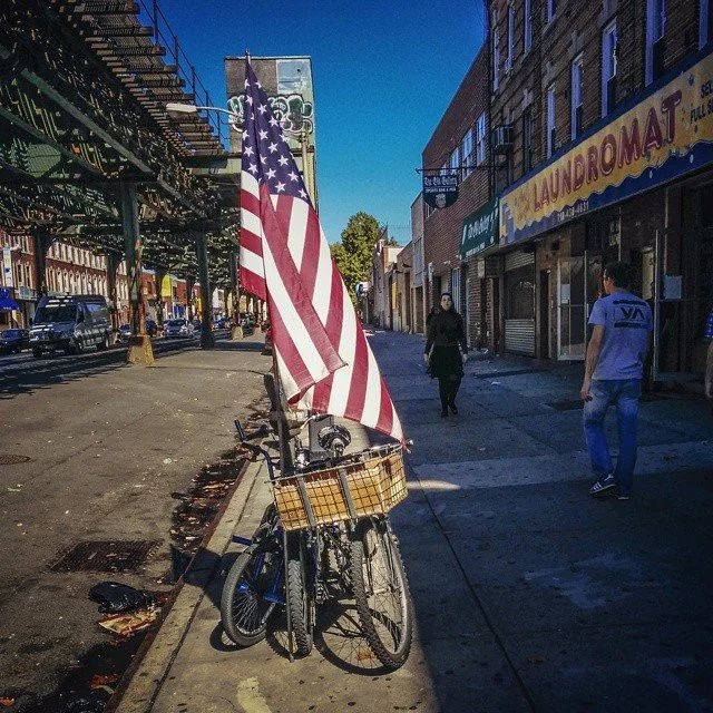 A bicycle with a basket parked on a city sidewalk with an American flag attached. Two people walk along the sidewalk, and storefronts and buildings line the street on a sunny day.
