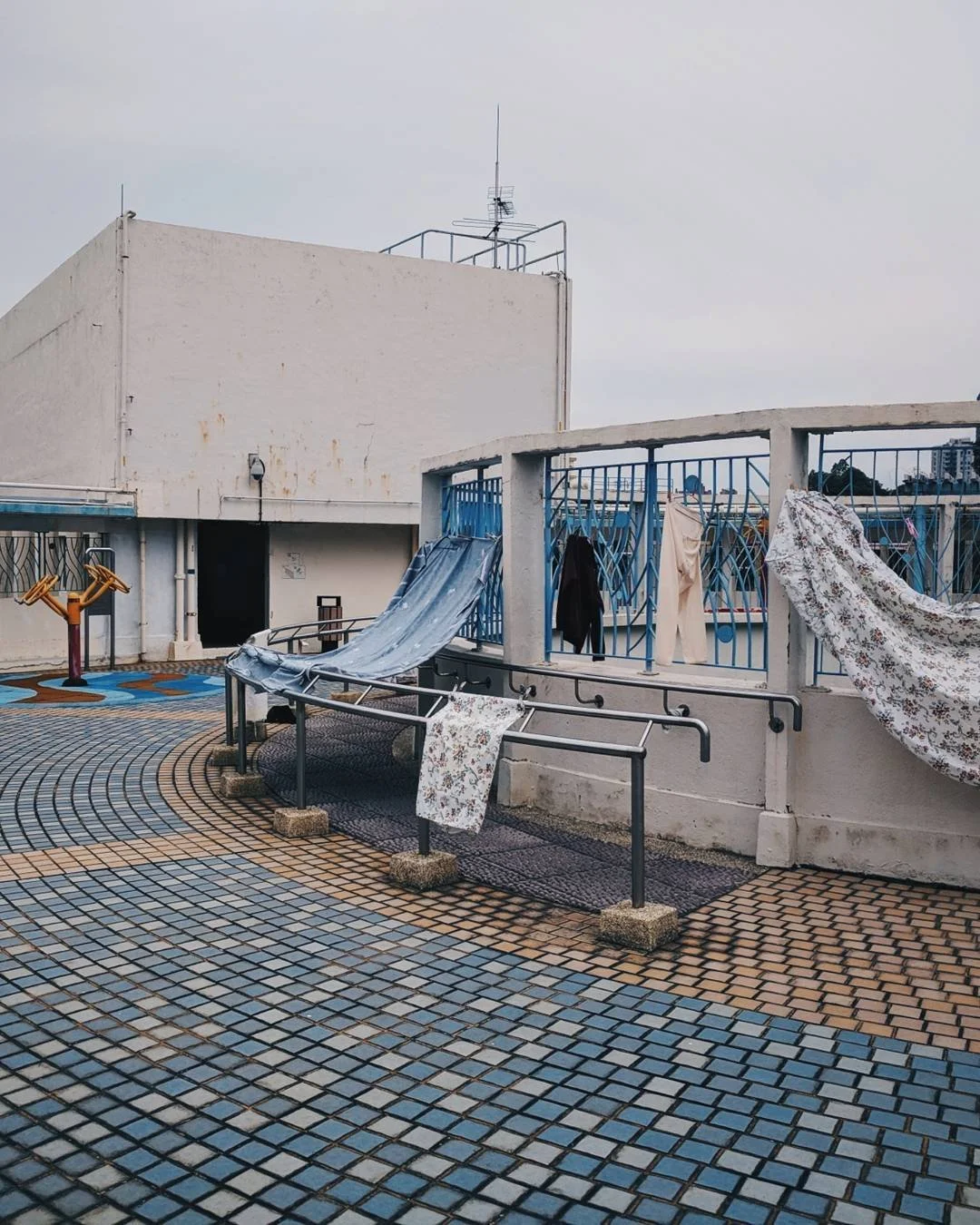 Outdoor rooftop with laundry hanging on a railing, including shirts, pants, and sheets, with a city skyline in the background and a cloudy sky.