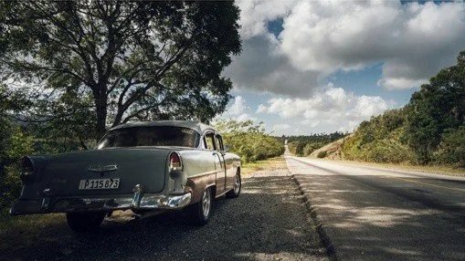 Vintage black car parked on the side of a rural road with trees and cloudy sky in the background.