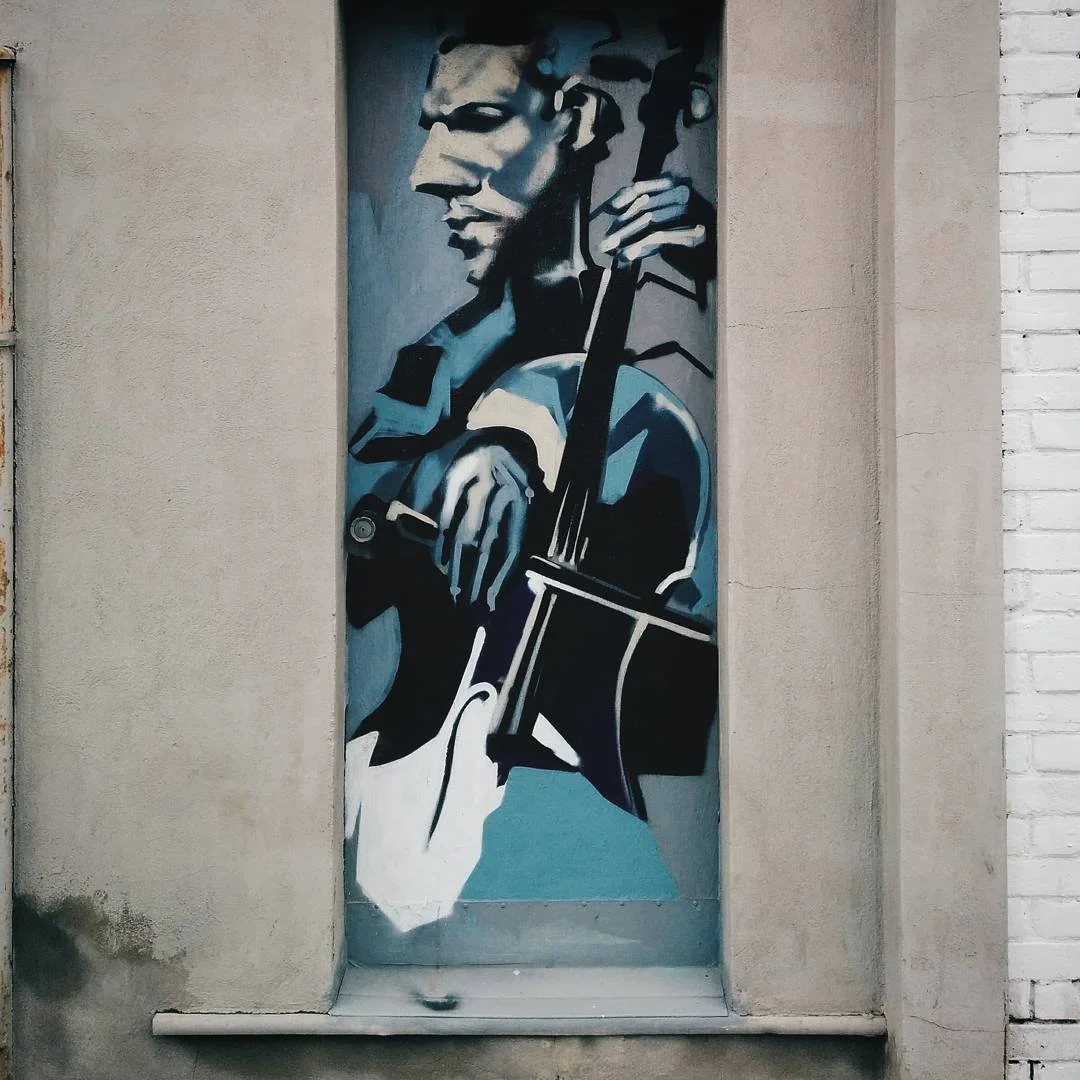 Street art mural of a person playing a cello, painted in black, white, and blue tones on a narrow wall in an urban setting.