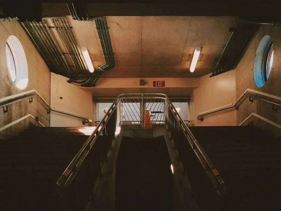 A staircase leading upwards with metal railings on both sides, viewed from below. The staircase is inside a building with concrete walls, round windows on both sides, and pipes along the ceiling. An illuminated exit sign is at the top of the staircase.