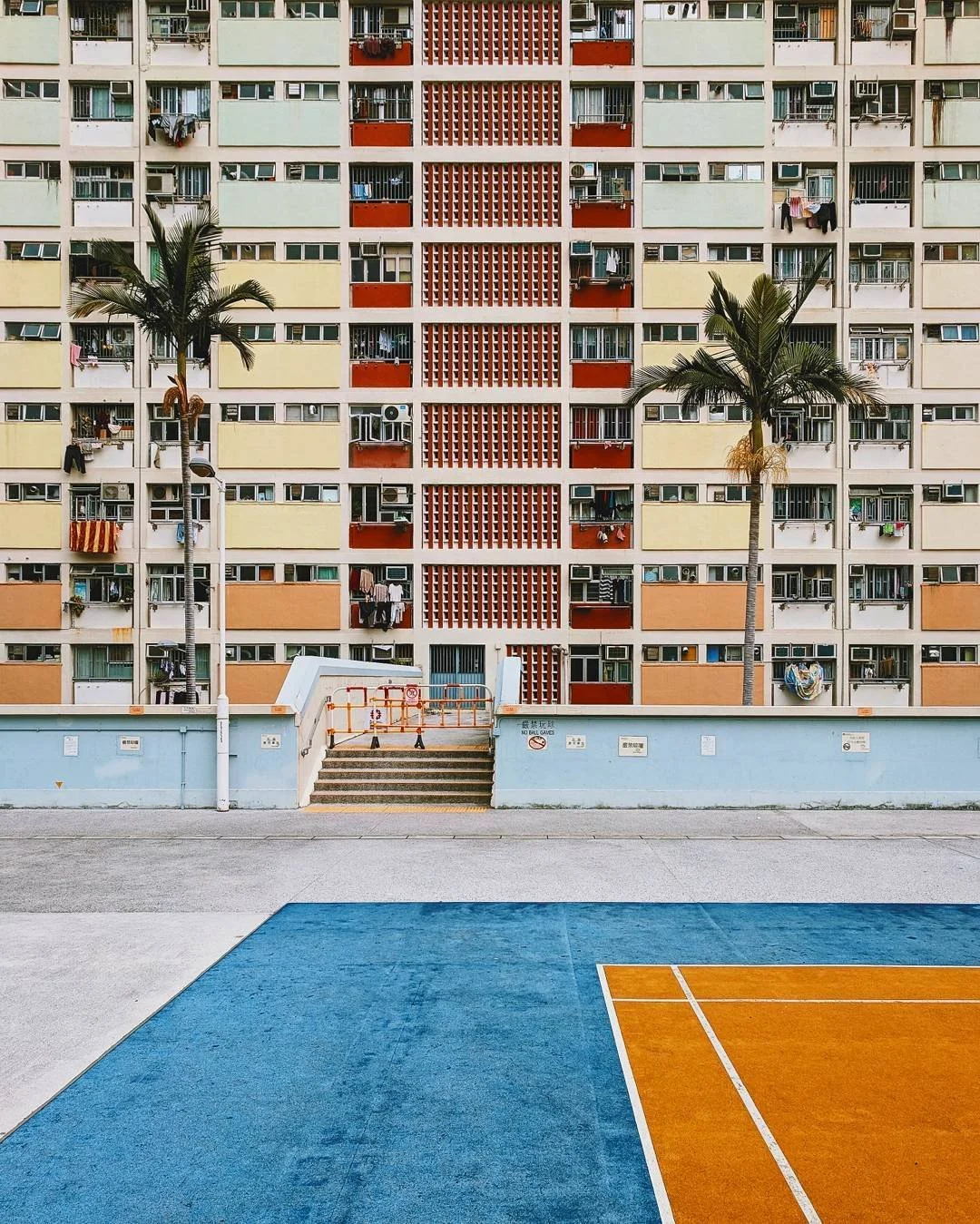Colorful apartment building with laundry hanging from windows, two palm trees in front, and a sports court with blue and orange sections in the foreground.