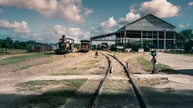 A vintage steam locomotive and other train cars on railroad tracks in front of a large open-sided shed, with a blue sky and scattered clouds overhead.