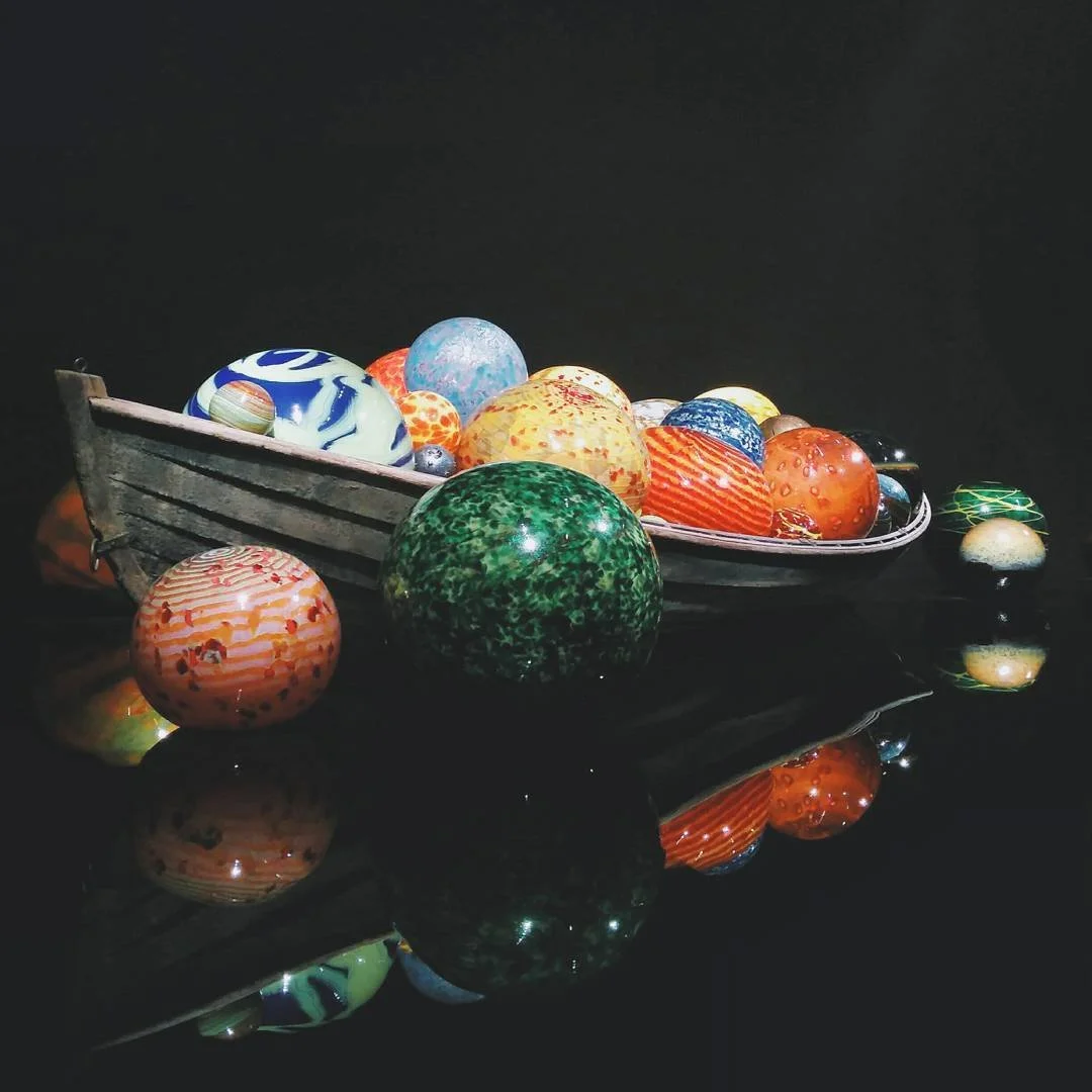 A boat-shaped display filled with colorful glass marbles and reflective glass balls, with some marbles and balls lying outside the boat, on a reflective surface.