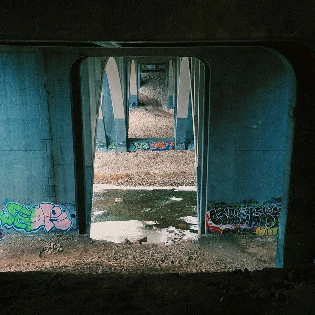 Under a bridge with graffiti, gravel and dirt on the ground, multiple arches recede into the distance.