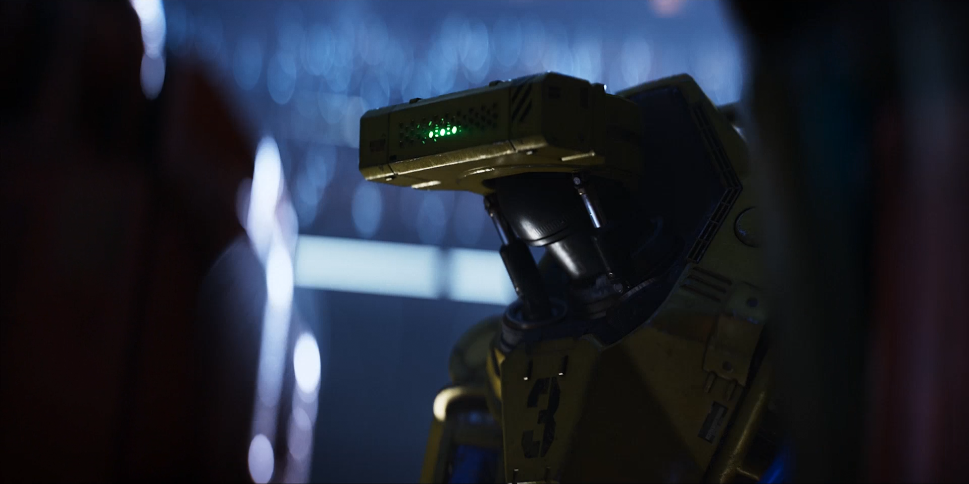A yellow robot with a black head and green lights on its forehead, standing in a dark environment with blurred lights in the background.