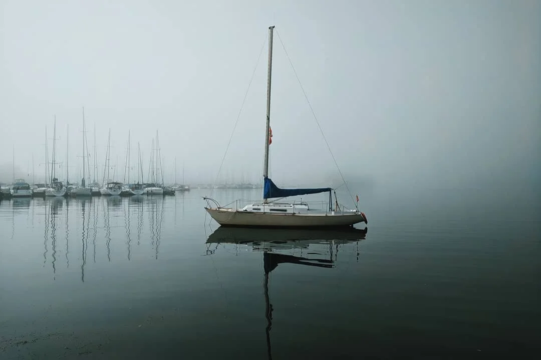 A sailboat floating on calm water with foggy background and other boats in the distance.