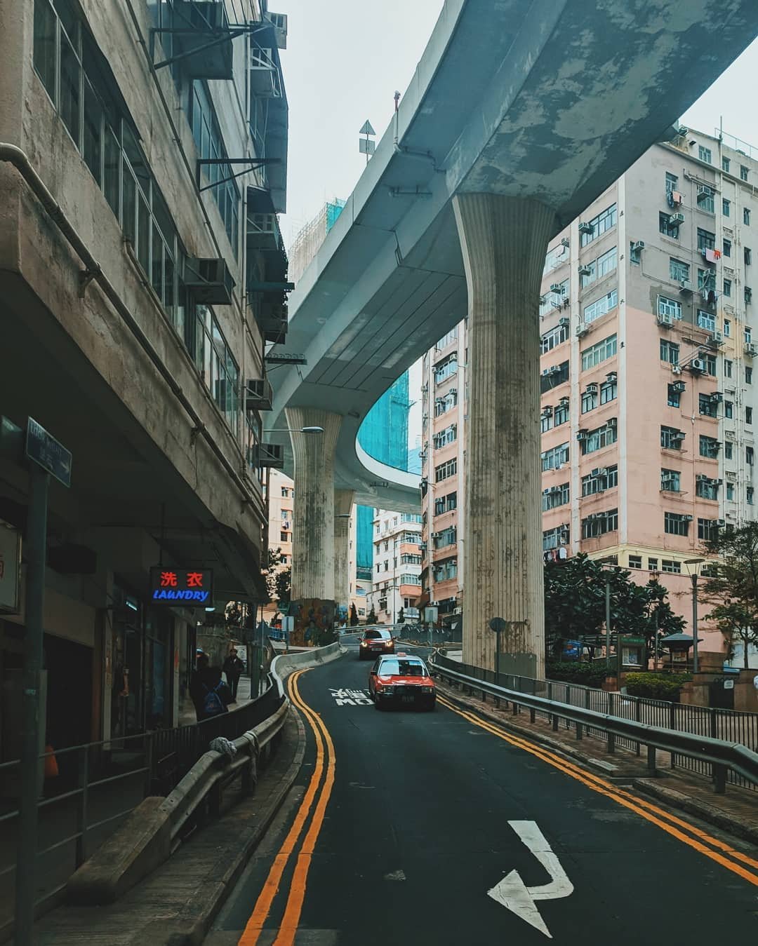 City street with elevated highway or train track overhead, surrounded by tall residential buildings; cars on curved road with yellow lines, a neon laundry sign on building, and pedestrians.