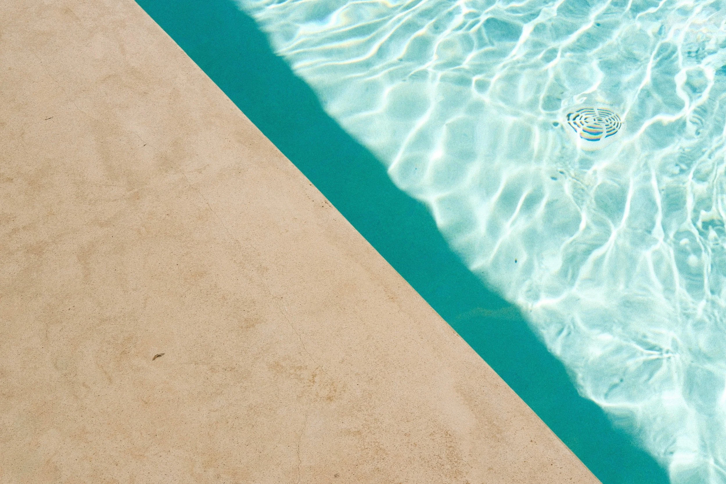 Close-up of a swimming pool's edge with clear blue water and sunlight reflections, showing a drain at the bottom.