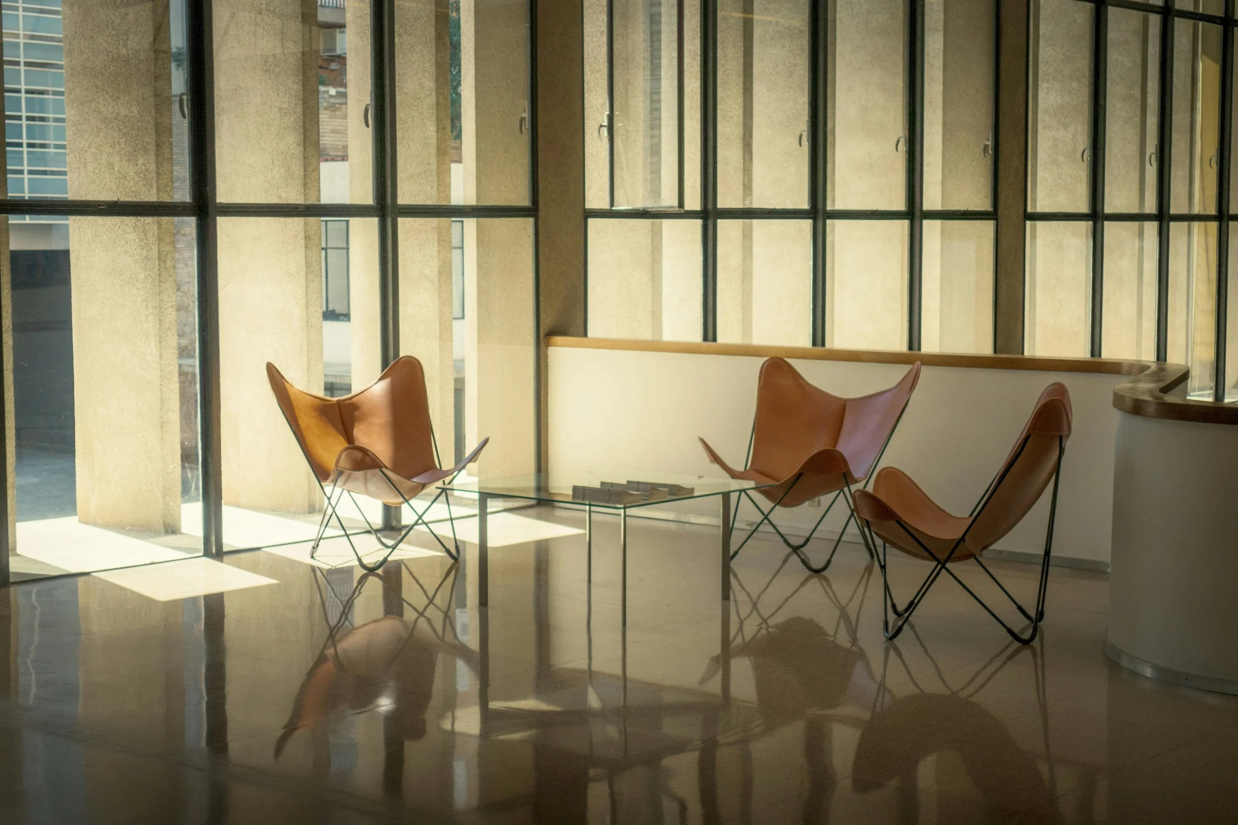A modern lounge area with three brown leather chairs around a glass coffee table, set against large windows with split panes letting in natural light.