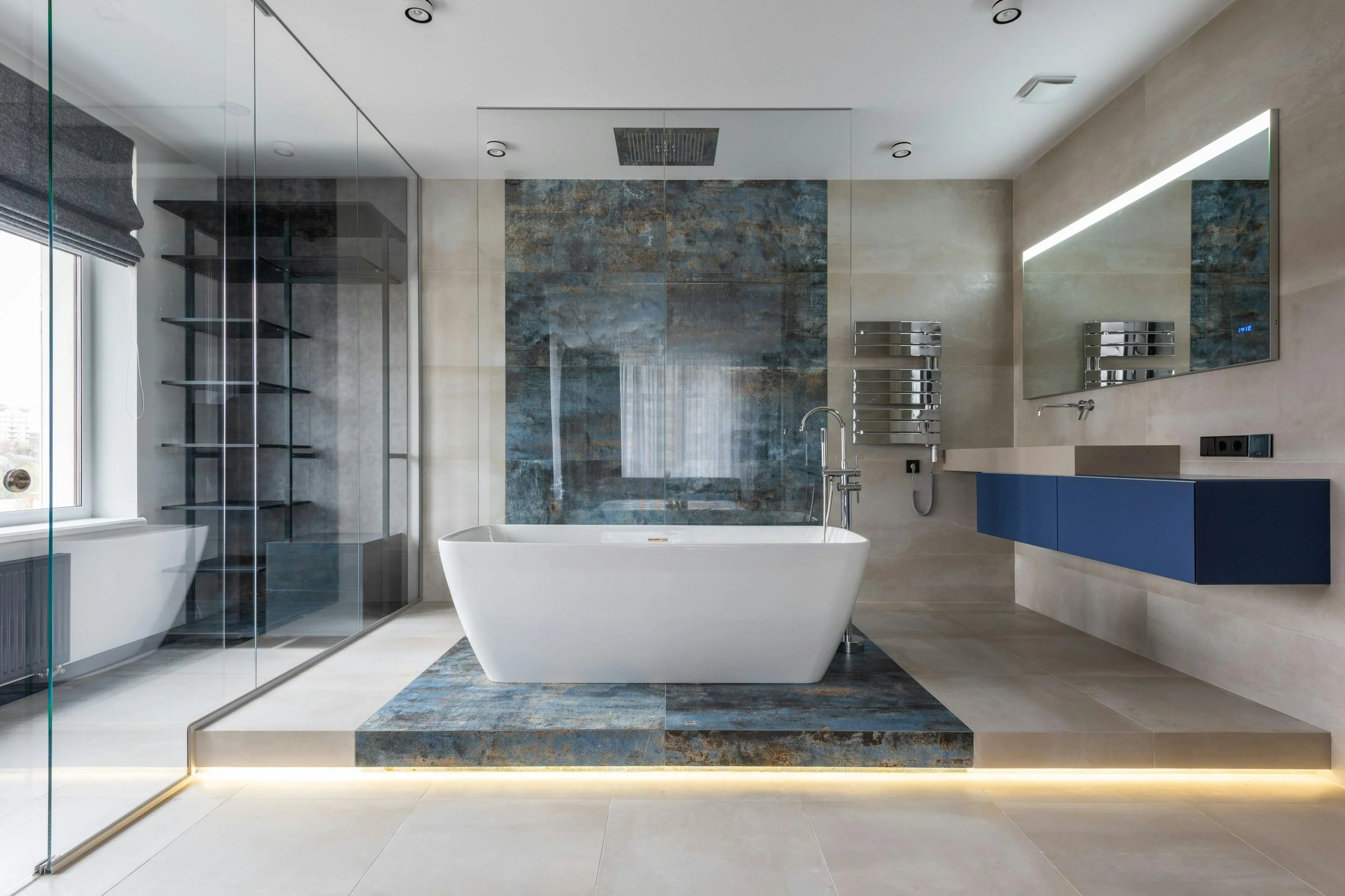 Modern bathroom with white bathtub on a raised stone platform, blue vanity, large mirror, glass shower enclosure, and window with blinds.