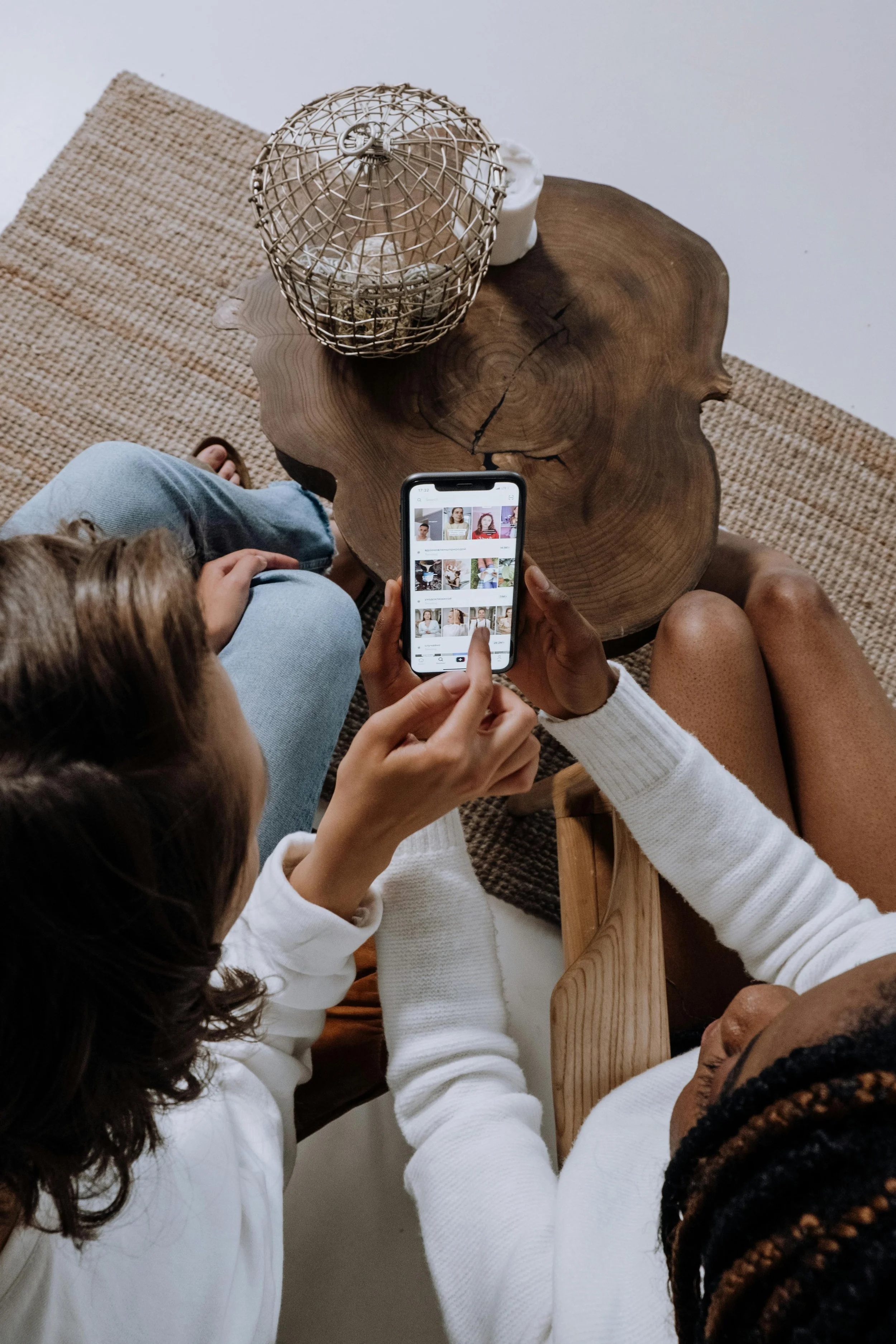 Two women sitting on a woven rug, looking at a smartphone together, with a wooden coffee table with decorative items on it nearby.