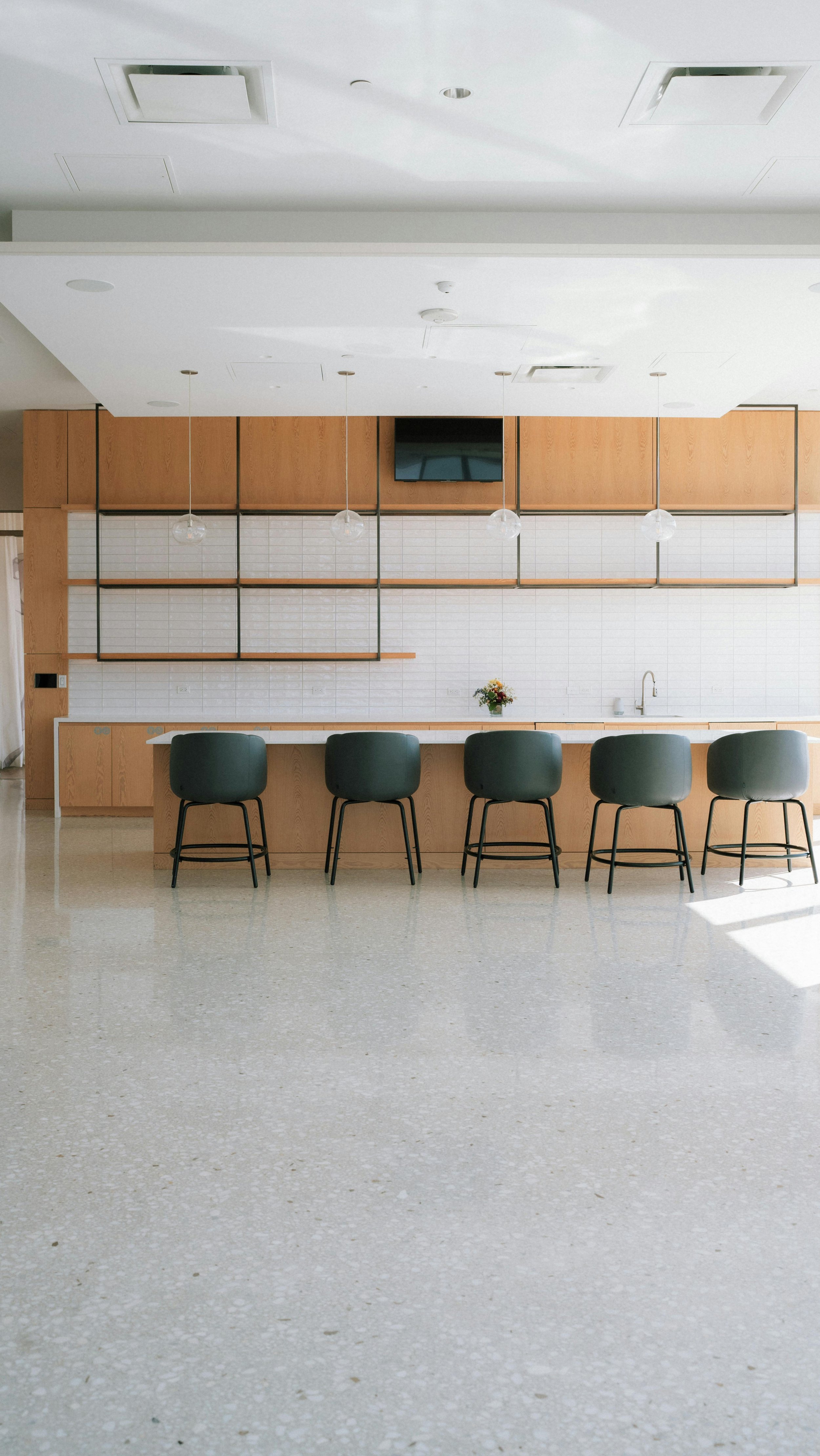 Modern kitchen area with a long countertop, black bar stools, wooden cabinets, white tiled backsplash, a vase with flowers, and hanging pendant lights.