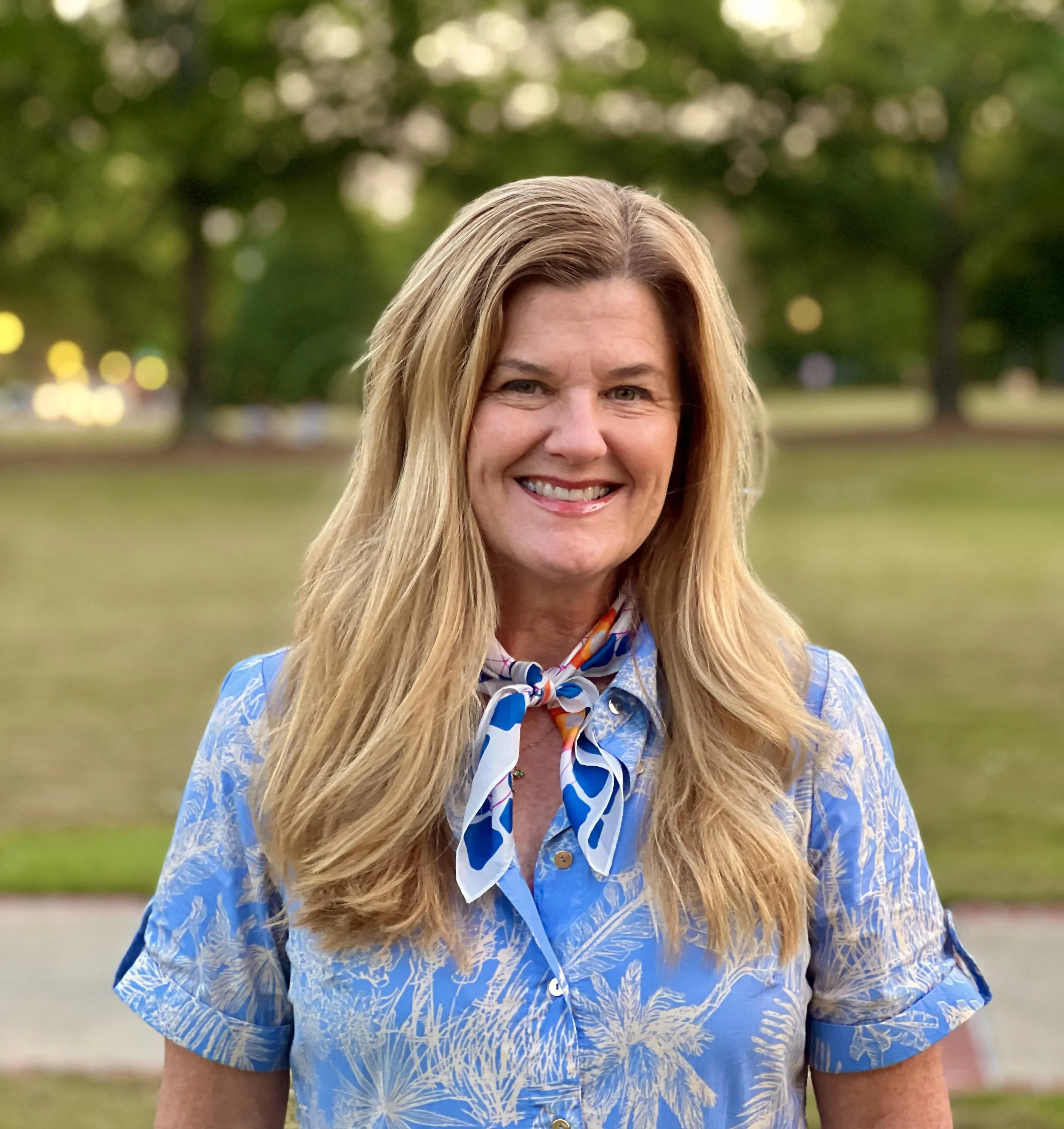 A woman smiling outdoors in a park, wearing a blue patterned shirt and a colorful scarf around her neck, with trees and blurred lights in the background.