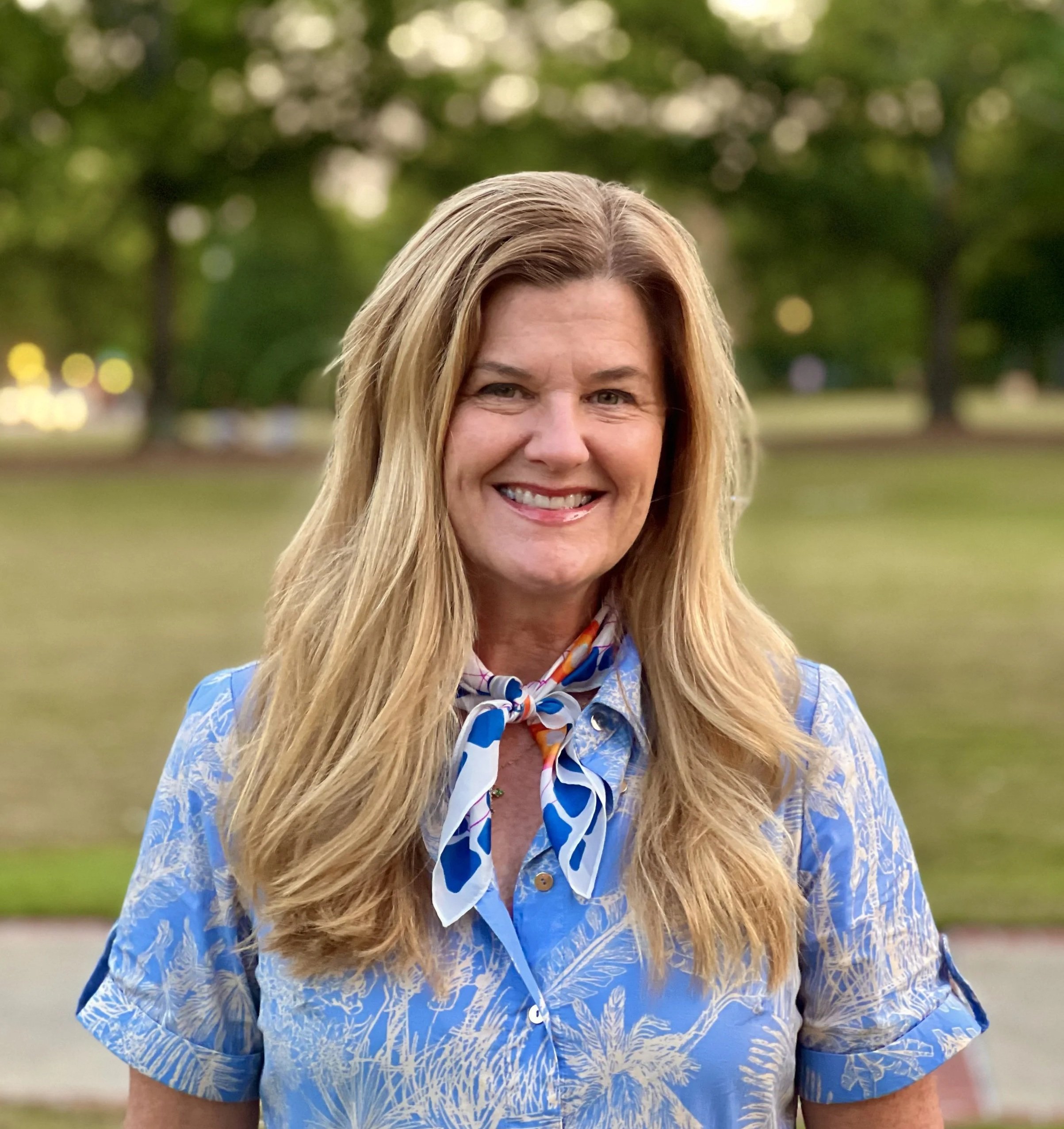 A woman smiling outdoors in a park, wearing a blue patterned shirt and a colorful scarf around her neck, with trees and blurred lights in the background.