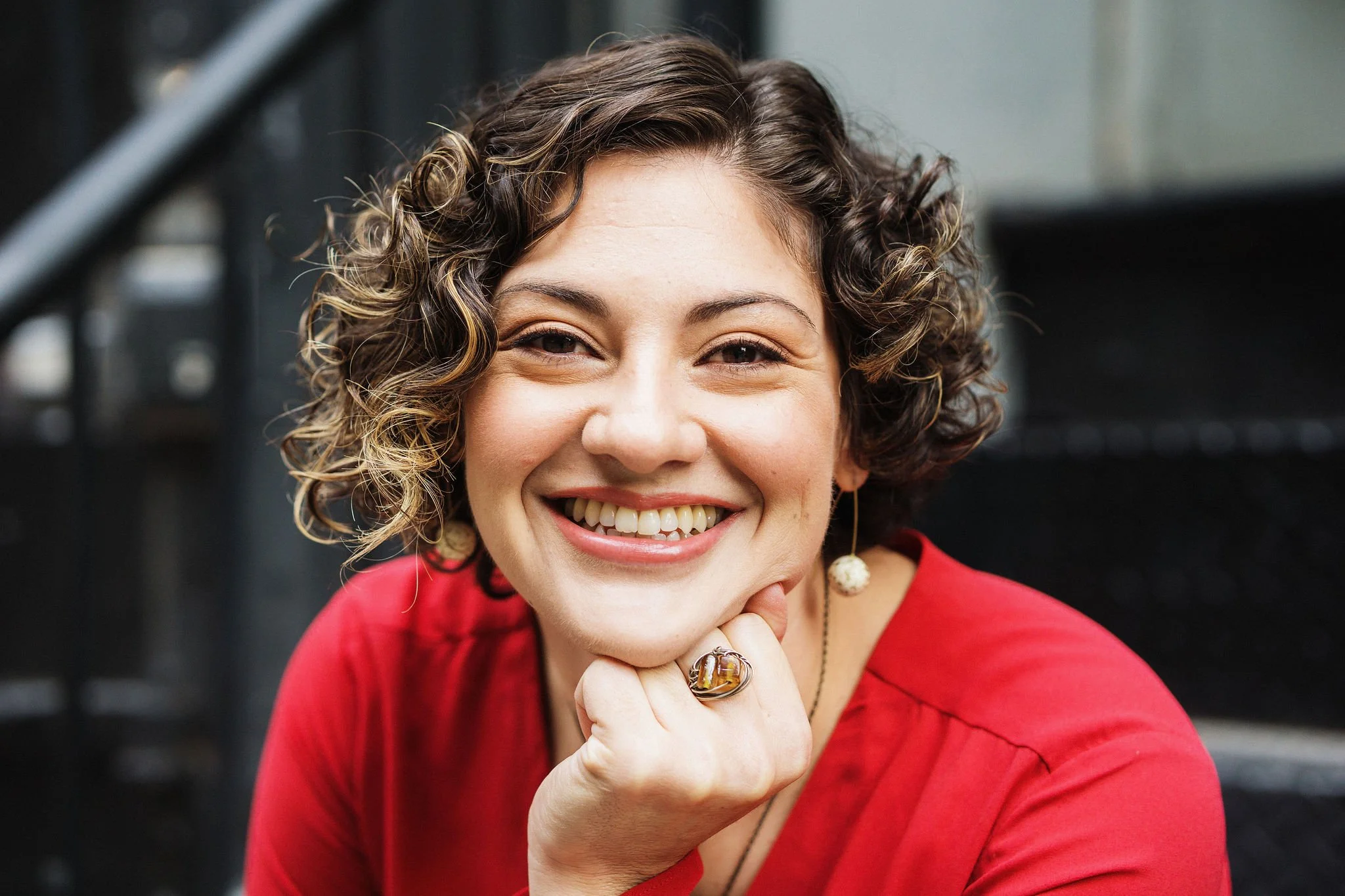 A white woman with brown curly hair and brown eyes, wearing a coral-colored blouse, smiles into the camera