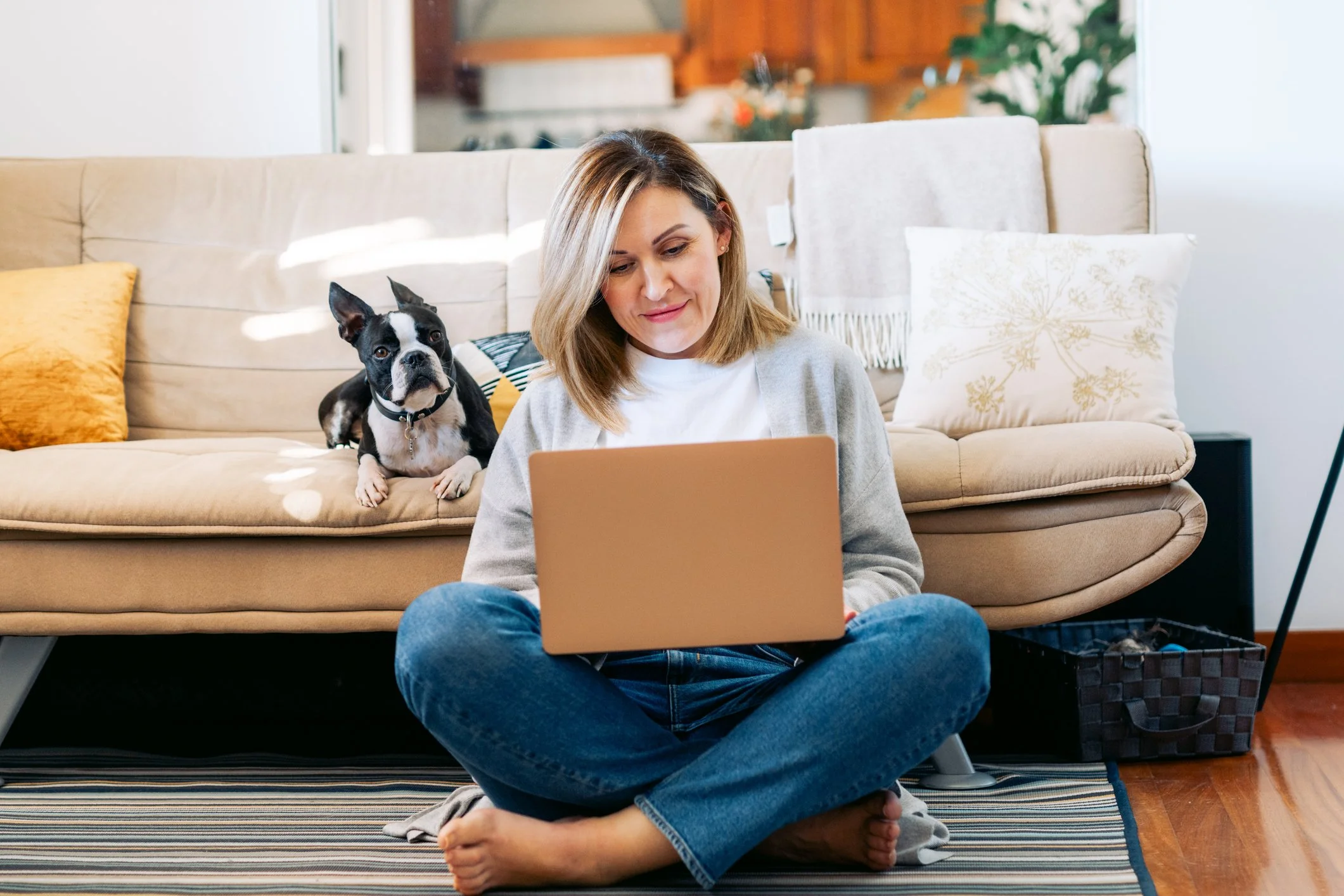 A woman working on a laptop while her Boston terrier relaxes on the couch, embodying balance and self-care promoted in online therapy sessions.
