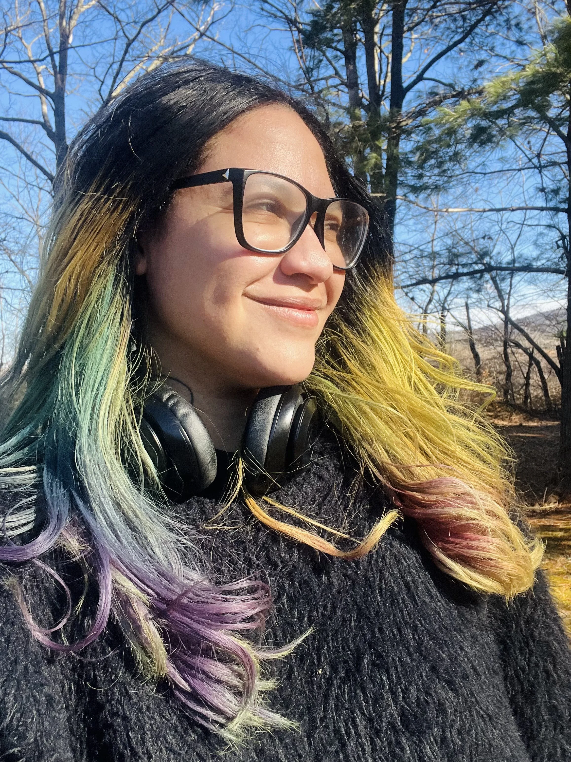 Elexis Trinity has light brown skin with black hair that is dyed with blue, purple and pink. They are smiling in the sun wearing glasses