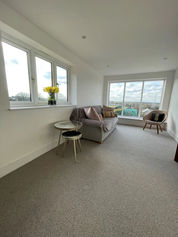 A bright room with large windows, a gray sofa with cushions, a small round side table with a black stool, a beige armchair, and a view of the outdoors, including cloudy skies and distant buildings.