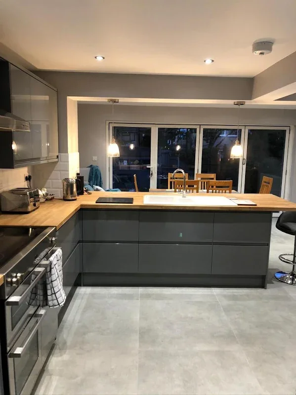 Modern kitchen with gray cabinets, wooden countertop, stainless steel appliances, and a sink. Overhead lighting and a dining area with wooden table and chairs visible through large sliding glass doors.