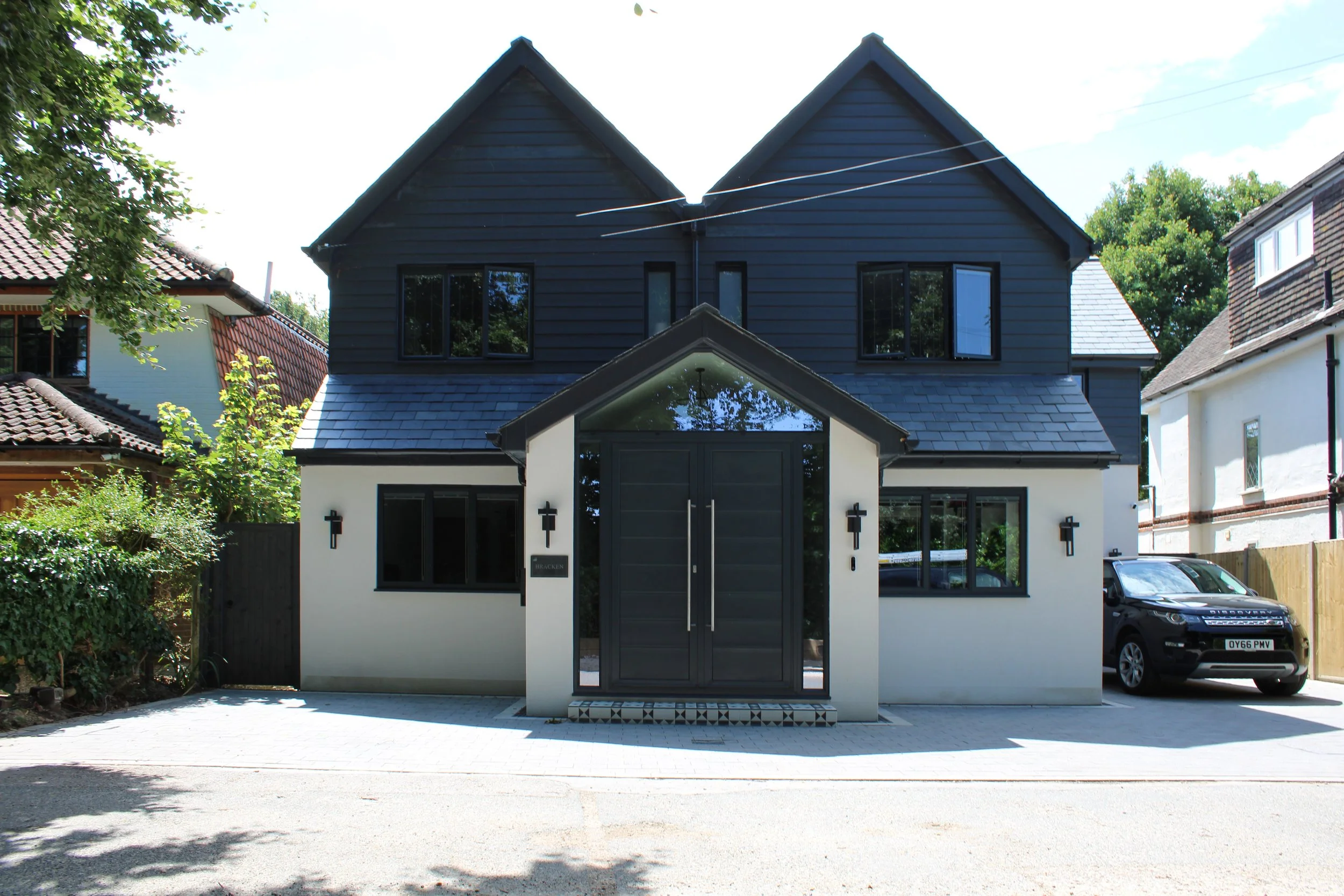 Modern two-story house with black and white exterior, black front door, black window frames, a driveway with a black car parked, surrounded by greenery and neighboring houses.