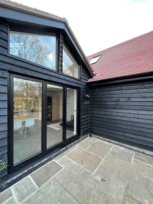 Black wooden house with large glass sliding doors and windows, a red-tiled roof, and a stone patio outside.