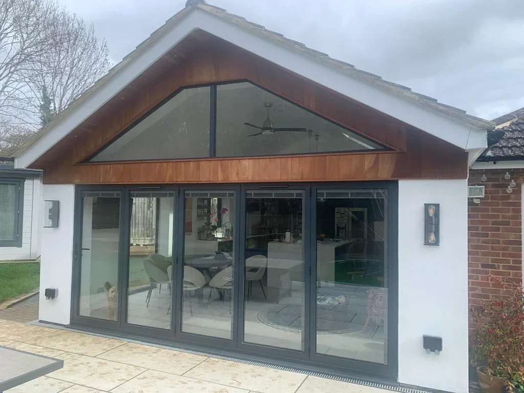 Modern house with large glass sliding doors and a triangular window on the gable end, showing indoor dining and living areas, outside patio, and a small dog on the patio.