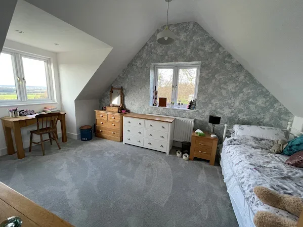 Bedroom with sloped ceiling, two windows, a packed bed, dresser, side table, desk, and chair.