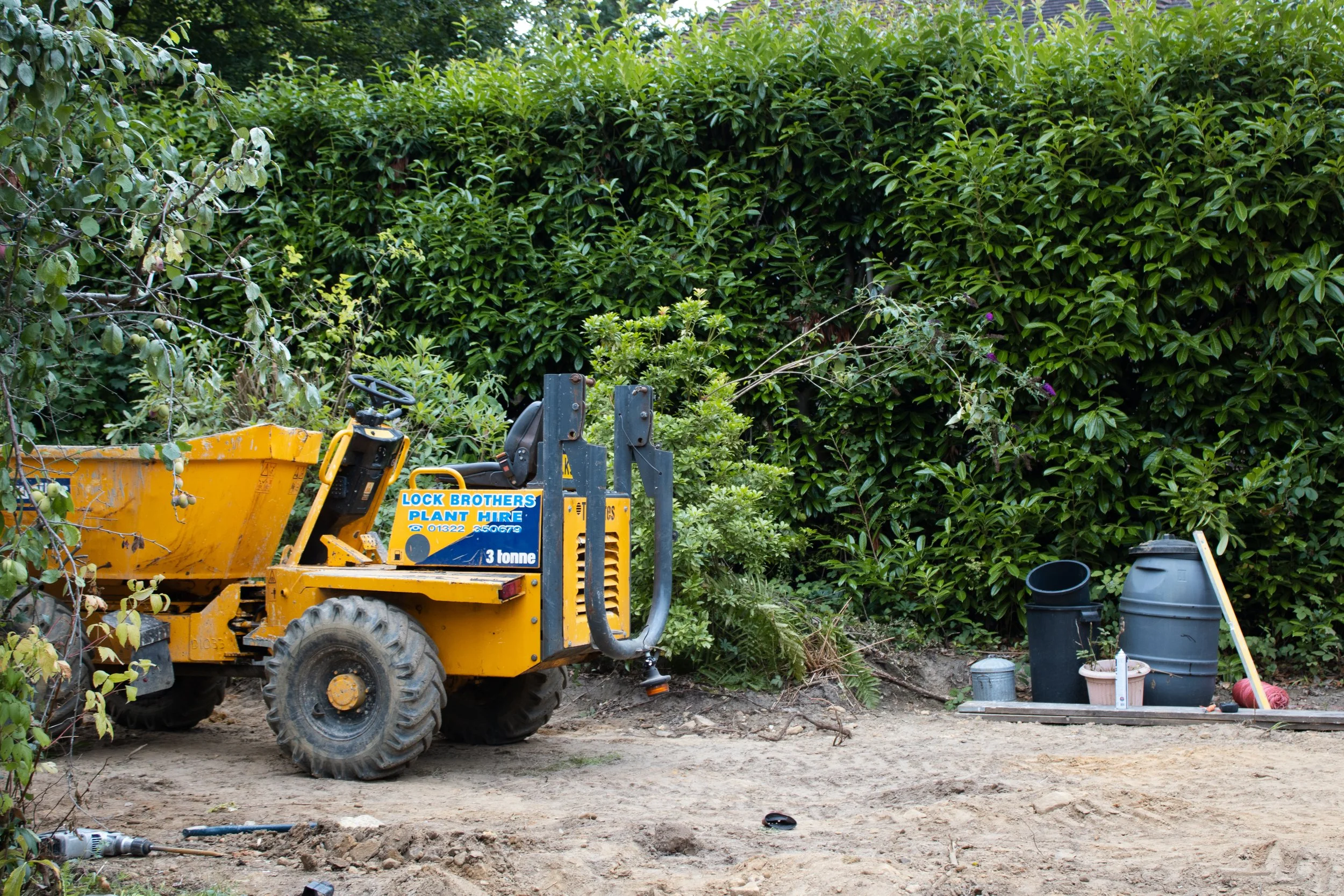 A small yellow construction vehicle parked on a dirt surface in front of a thick green hedge. There are various gardening supplies, including buckets, a trash can, and a watering can, on the right side of the image.