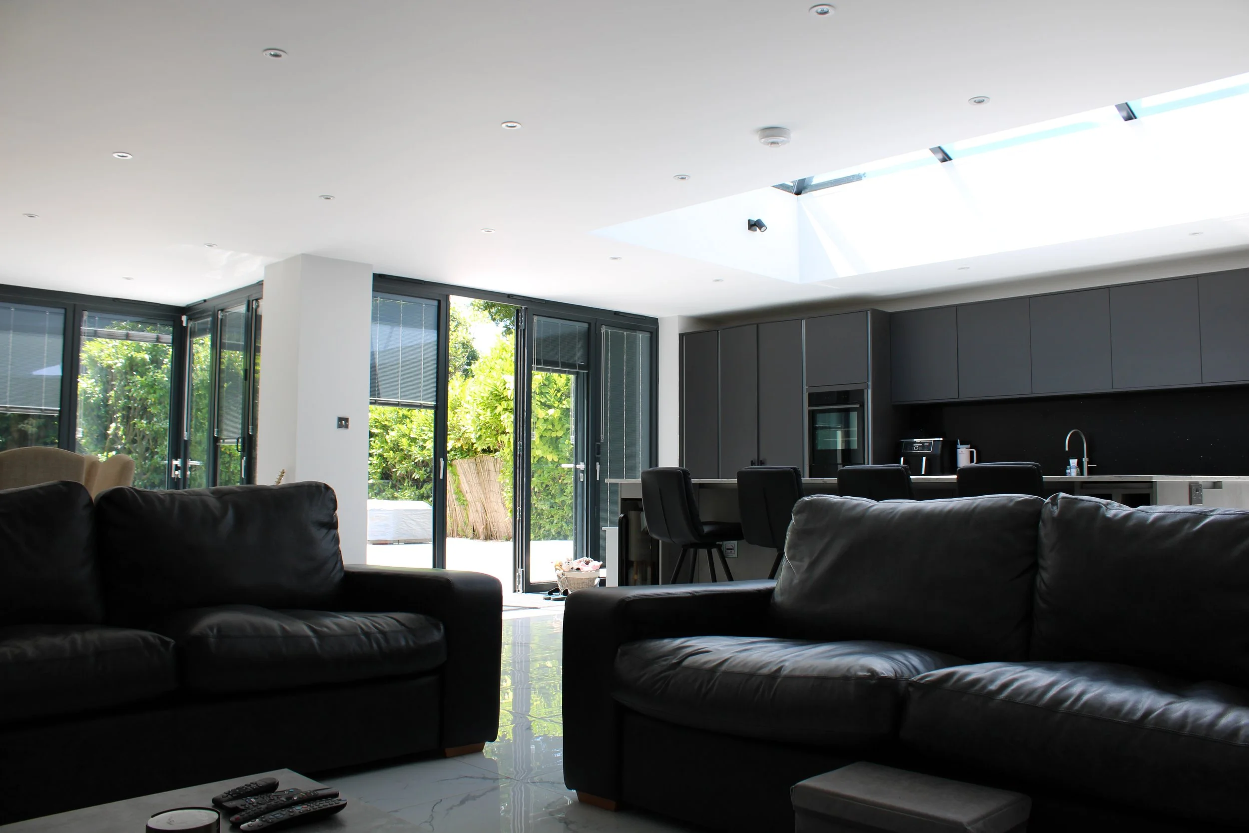 Modern living room with black leather sofas, large glass doors leading to a garden, and a kitchen with dark cabinets, barstools, and a skylight.