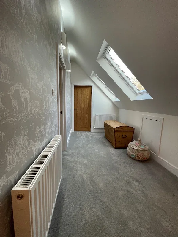 An attic room with skylight windows, a wooden chest, a wicker basket, a radiator, and gray carpeting.