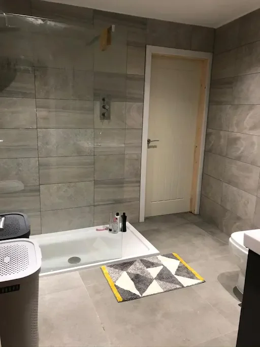 Bathroom with gray tiled walls and floor, a door, shower area with several bottles, laundry basket, and a small rug