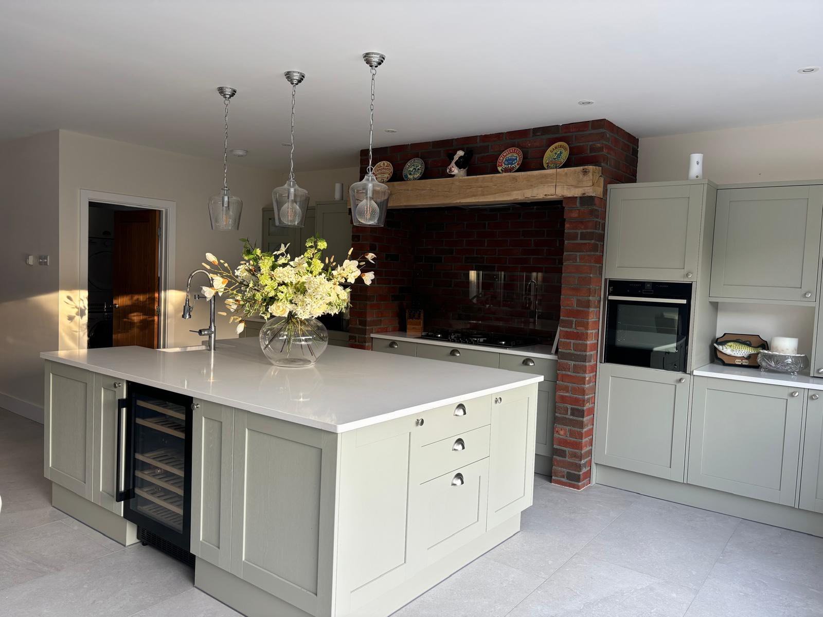 Modern kitchen with light green cabinets, white countertop island with a vase of flowers, brick fireplace with decorative plates on the mantle, gray tiled floor, and pendant lighting.
