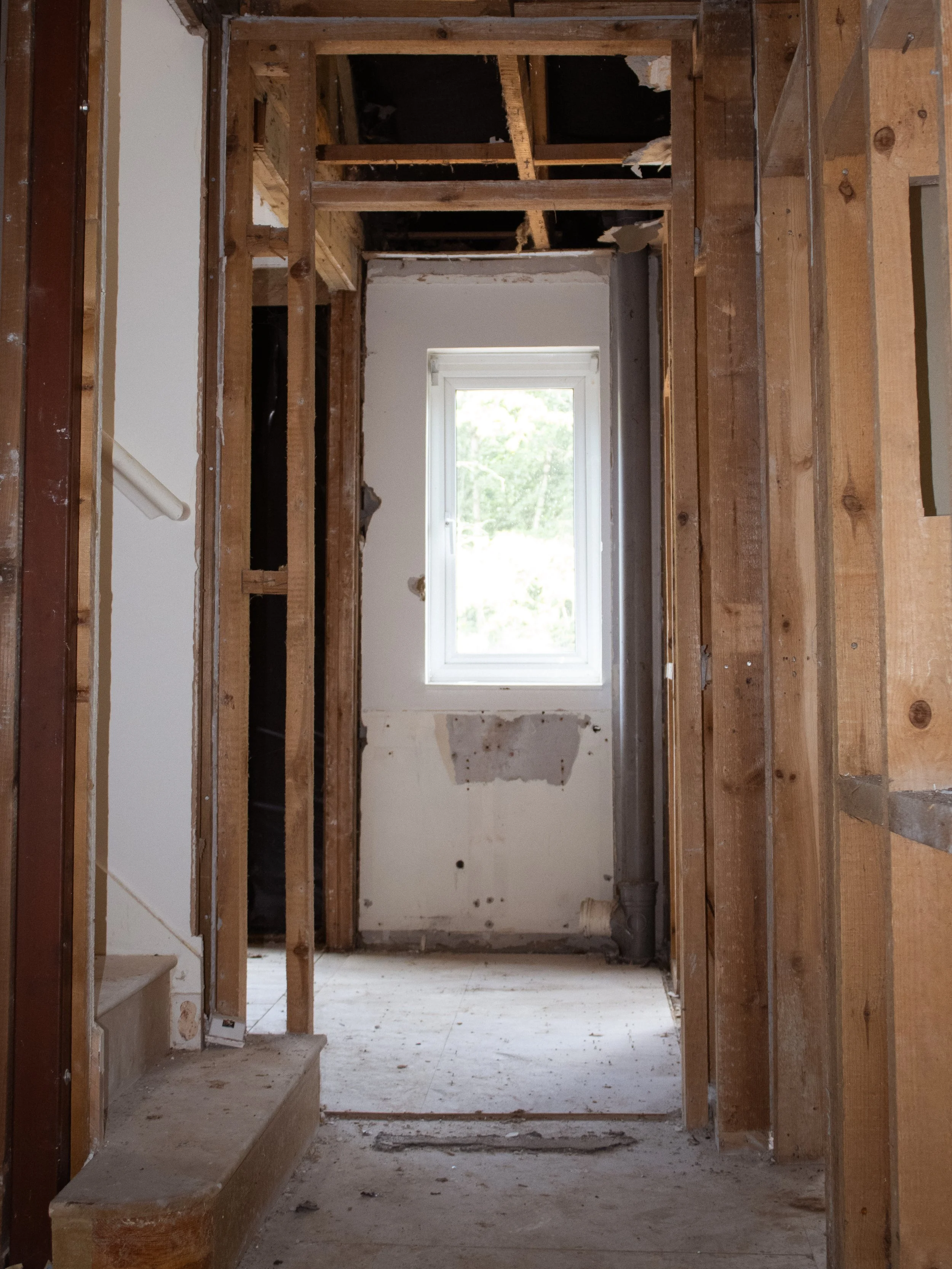 Interior of a house under construction, showing exposed wooden framing, a window at the end, and construction debris on the floor.