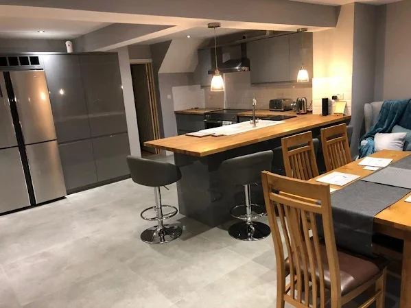 Modern kitchen with a breakfast bar, gray cabinets, stainless steel refrigerator, hanging pendant lights, wooden dining table with chairs, and gray tiled flooring.