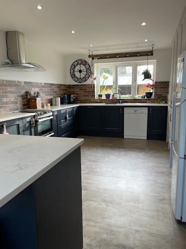 Modern kitchen with dark blue cabinets, a brick backsplash, a white countertop, and a window with plants on the sill.