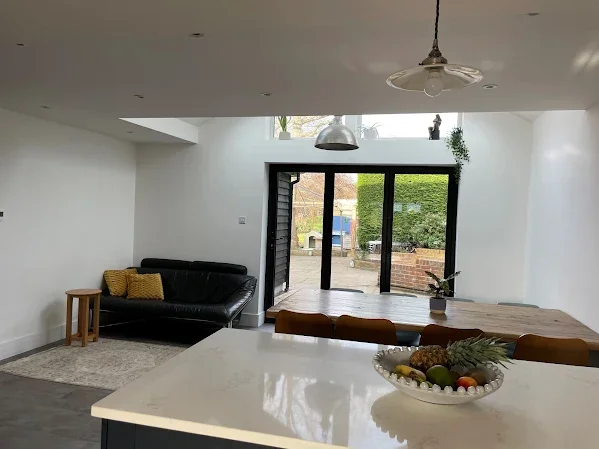 Modern living room with black leather sofa, wood side table, dining table, fruit bowl, large glass sliding doors, and outdoor garden view