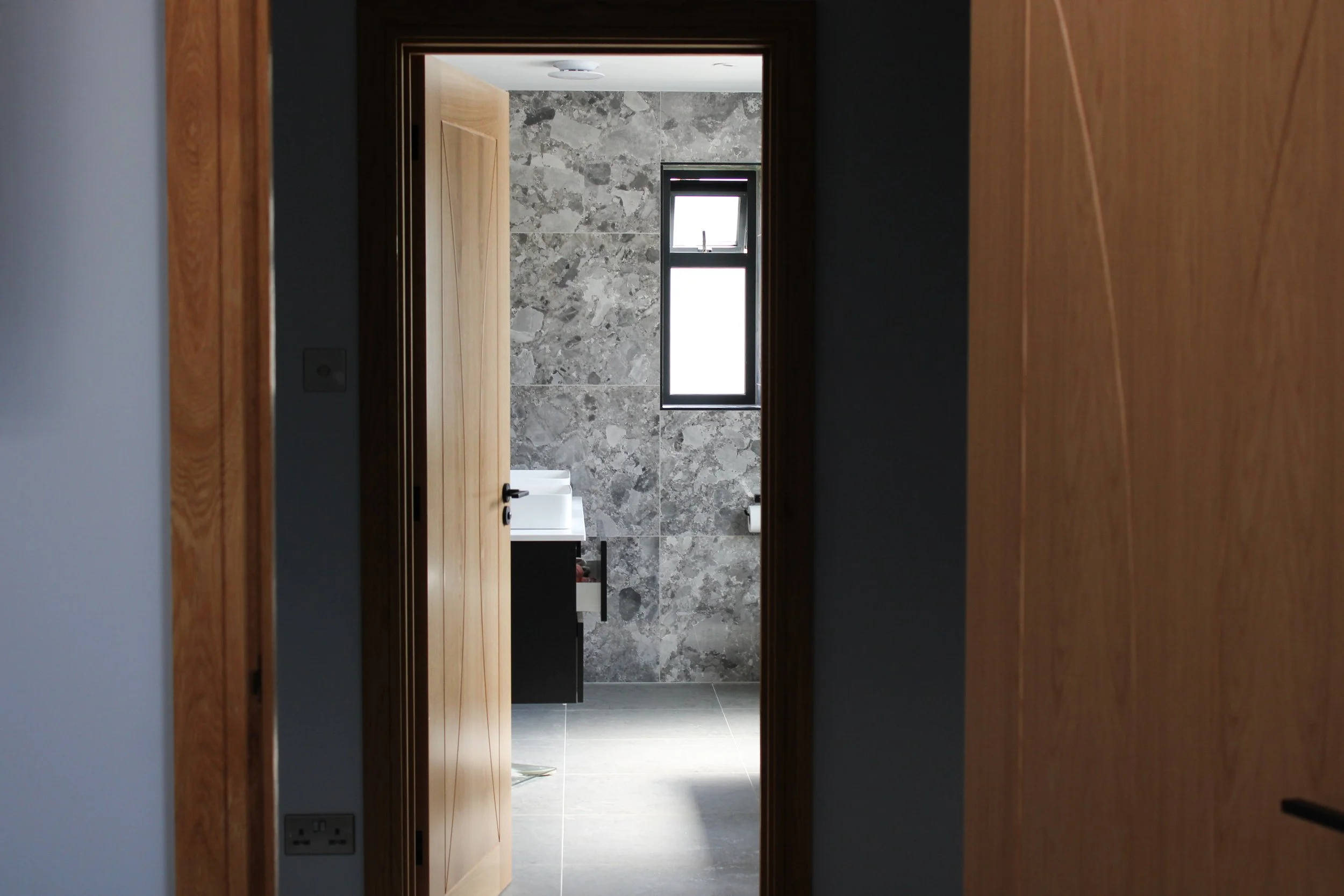 View into a modern bathroom through an open wooden door, showing a gray stone tiled wall, a small window, and a black and white vanity.